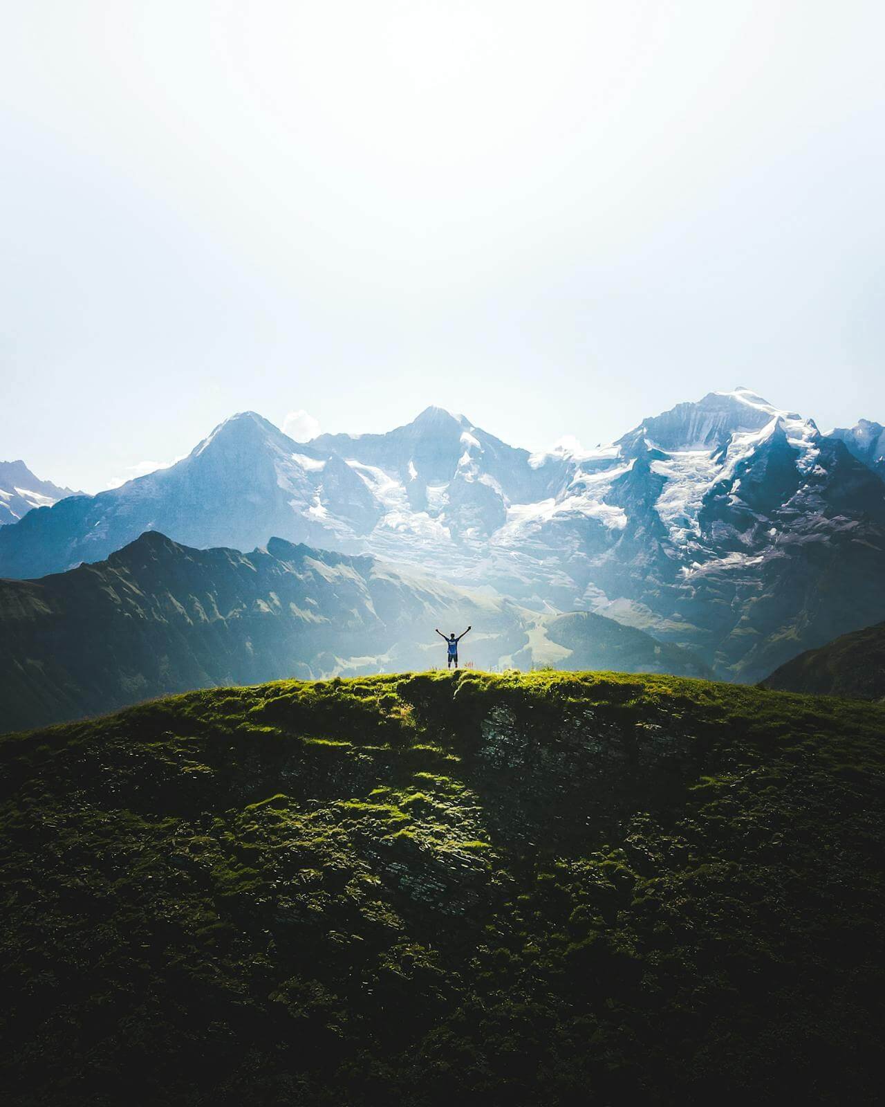 Žmogus pakeltomis rankomis stovi ant žalios kalvos prieš snieguotus kalnus – svetainės matomumo rezultatai PRIEŠ (be tekstų) ir PO (su SEO tekstais)